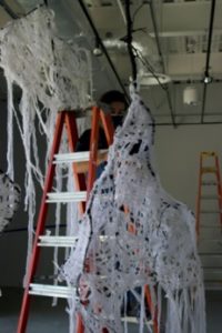 Image of sculpture consisting of large branches wrapped with white fabric, hanging from the gallery ceiling 