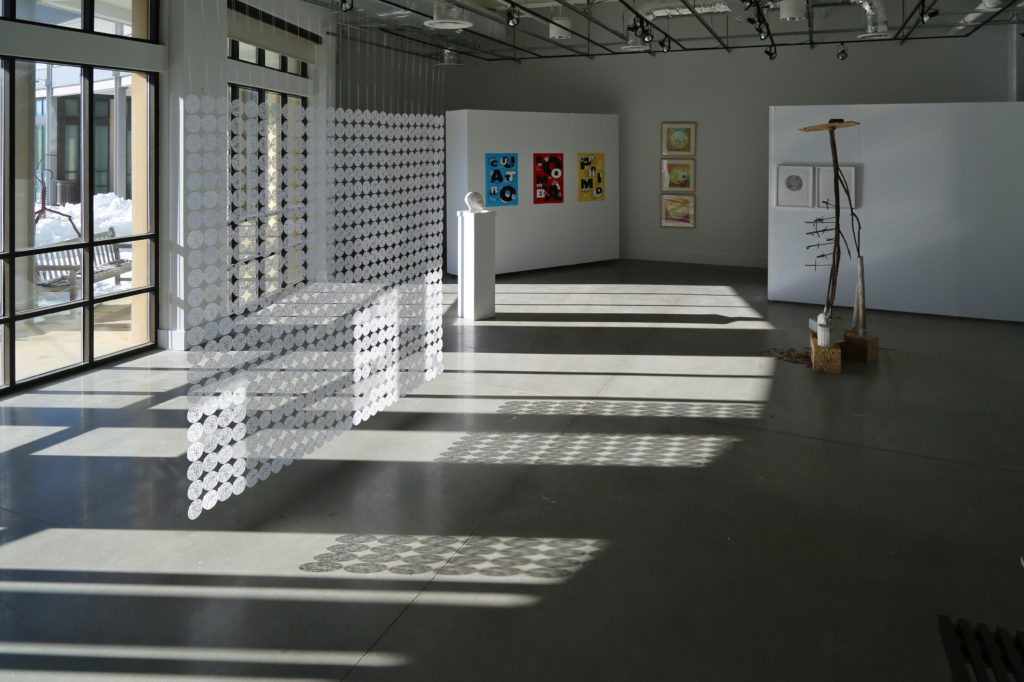 View of the exhibition in the gallery, with windows along the left side, light streaming in, and views of a few artworks 