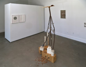 View of a tall sculpture made from branches as legs, on plywood boxes. There is a framed drawing of an eye opposite the sculpture.