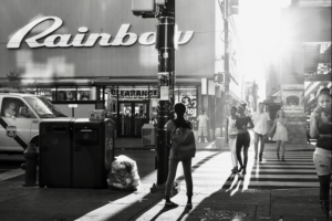 A black and white photo of a busy city street. Sunlight highlights the silhouette of a student facing away from the camera.